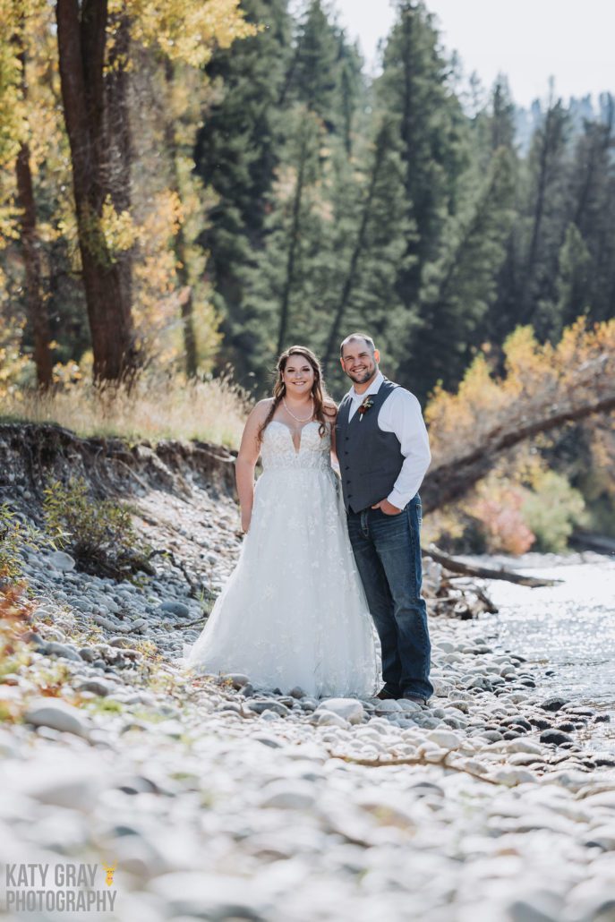 A bride and groom stand on the pebbled shore of the Snake River.