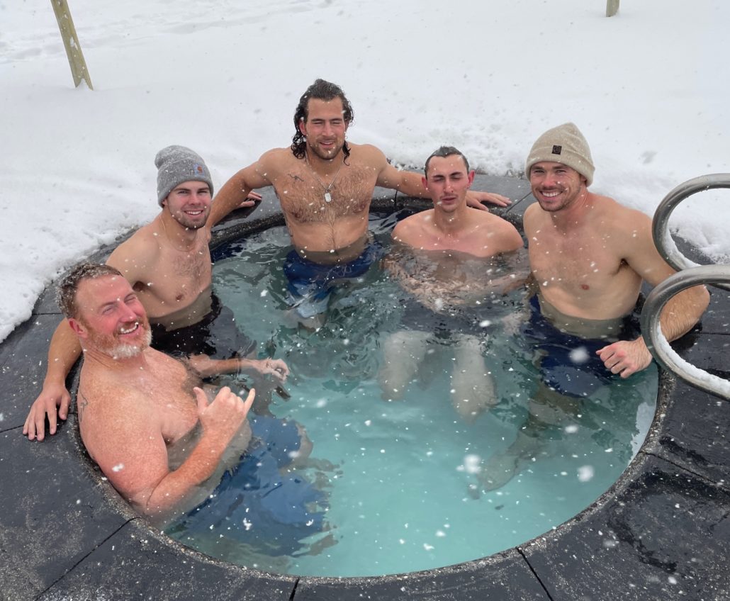 A group of five men stand in a cold plunge pool on a snowy day.