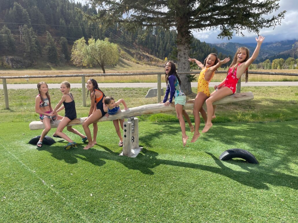 A ground of young girls in bathing suits play on a wooden see-saw.